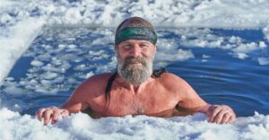 Ein Mann mit Bart taucht in einem mit Eis umgebenen Gewässer auf. Er trägt ein Stirnband und blickt ruhig zur Kamera. Die Umgebung ist winterlich und verschneit. die Wim Hof Atmung hilft, um besonders im Winter ein gutes Immunsystem zu habenn.
